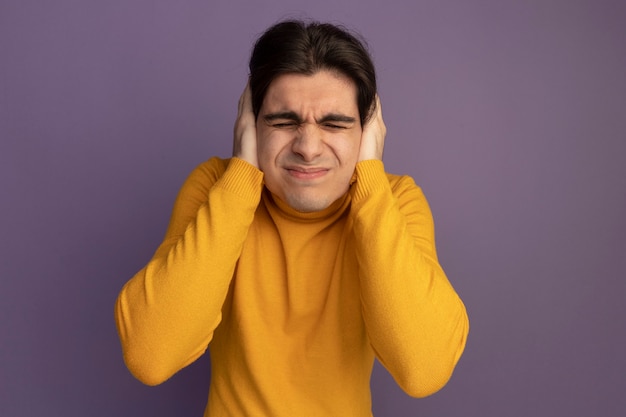 Insatisfecho con los ojos cerrados joven guapo con suéter de cuello alto amarillo poniendo las manos en las orejas aisladas en la pared púrpura