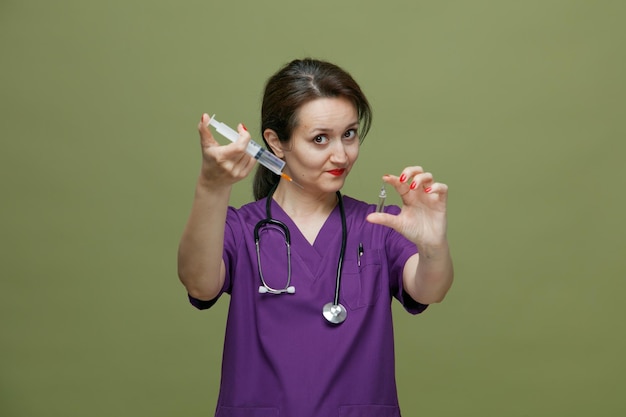 Foto gratuita insegura doctora de mediana edad con uniforme y estetoscopio alrededor del cuello mirando a la cámara estirando la jeringa con aguja y ampolla médica hacia la cámara aislada en el fondo verde oliva