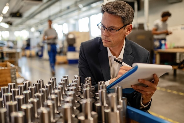 Inspector de control de calidad que escribe informes mientras analiza cilindros de varilla de acero fabricados en edificios industriales