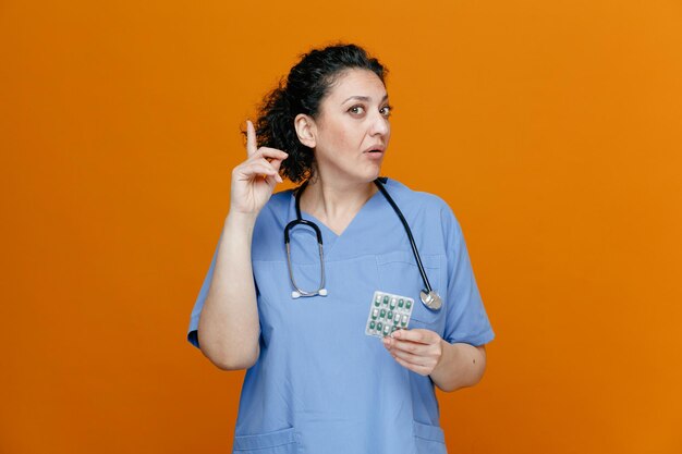 Inspirada doctora de mediana edad con uniforme y estetoscopio alrededor del cuello sosteniendo un paquete de pastillas mirando a la cámara apuntando hacia arriba aislada en el fondo naranja