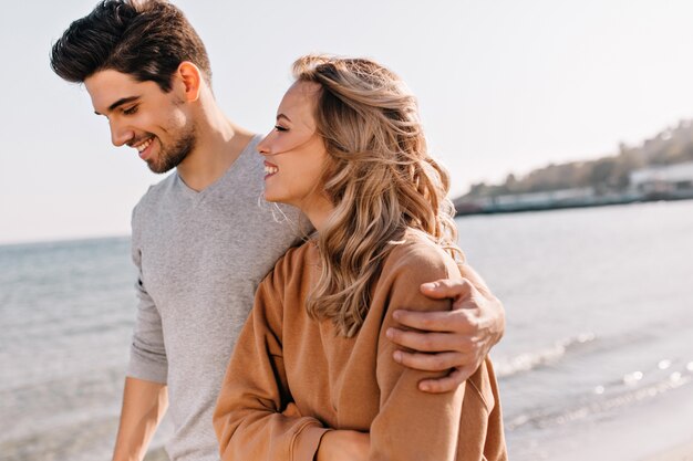 Inspirado joven abrazando a novia durante el paseo por la playa. Mujer rubia curiosa pasar el fin de semana en el mar.