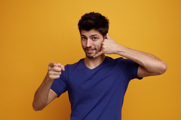 Inspirado joven apuesto mirando a la cámara haciendo un gesto de llamarme apuntando a la cámara con fondo amarillo