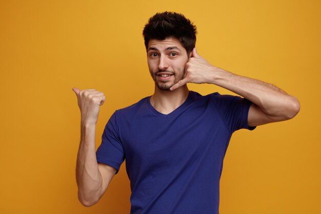 Inspirado joven apuesto mirando a la cámara haciendo un gesto de llamarme apuntando a un lado sobre fondo amarillo