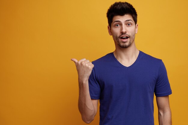 Inspirado joven guapo apuntando al lado mirando a la cámara sobre fondo amarillo con espacio de copia