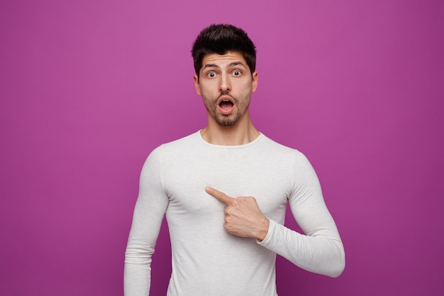 Inspirado joven guapo mirando a la cámara apuntándose a sí mismo sobre fondo púrpura