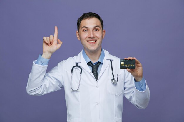 Inspirado joven médico masculino con bata médica y estetoscopio alrededor del cuello mirando a la cámara que muestra la tarjeta de crédito apuntando hacia arriba aislada en el fondo morado