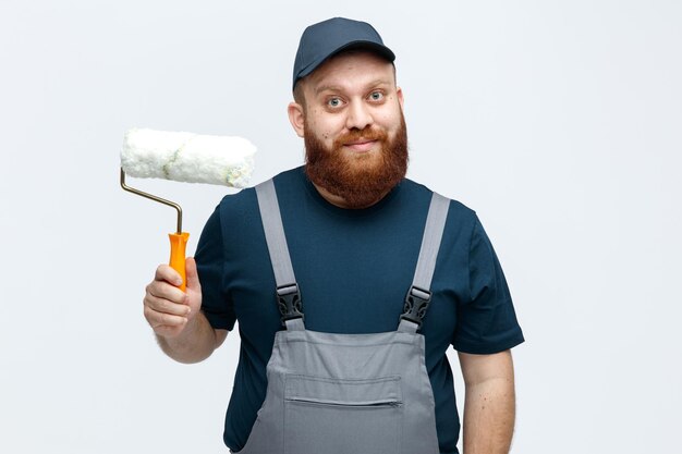 Inspirado joven trabajador de la construcción con gorra y uniforme sosteniendo un rodillo de pintura mirando a la cámara aislada en el fondo blanco