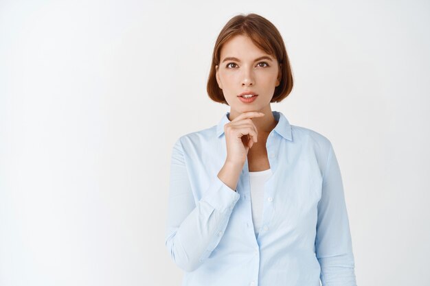 Inspírate. Retrato de joven directora directora que mira pensativa, tiene una idea, toca la barbilla y reflexiona sobre el plan, de pie en la pared blanca