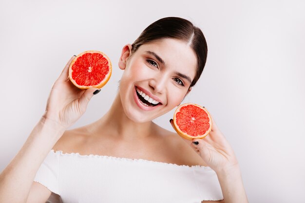 Instantánea de una joven de ojos grises con una sonrisa blanca como la nieve que muestra pomelos jugosos y saludables en la pared blanca.