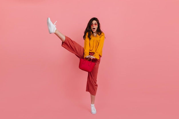 Instantánea de una morena alegre con pantalones rosas levantando la pierna en alto Dama con un elegante atuendo posando con un bolso en un fondo aislado