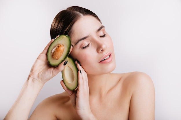 Instantánea de niña sin maquillaje en pared blanca. Señora con los ojos cerrados mantiene el aguacate saludable.