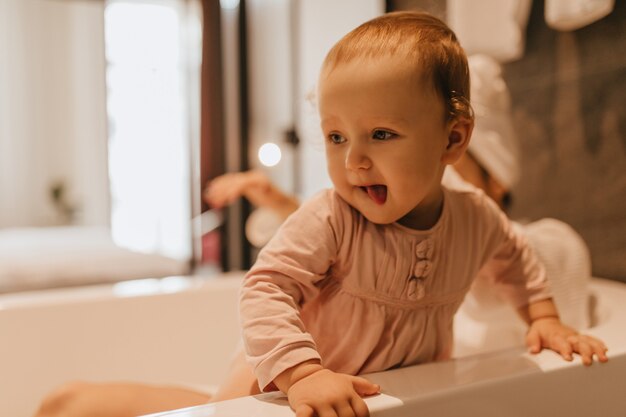 Instantánea de niño rubio curioso en blusa rosa estudiando objetos con interés en el baño.