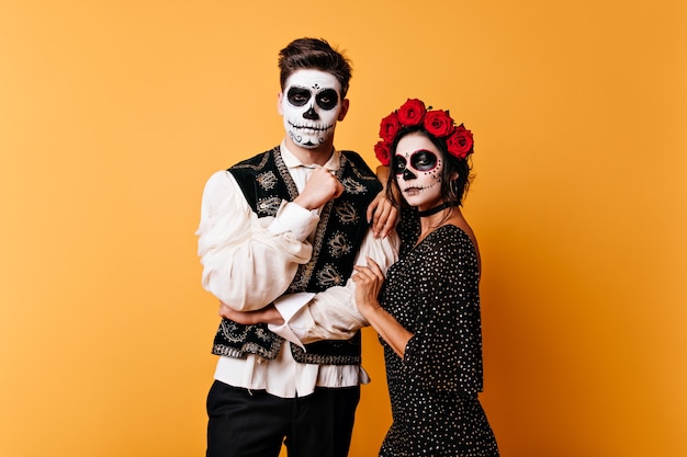 Instantánea de pareja con arte facial para el día de todos los muertos. Mujer joven con corona de rosas abraza a chico de pelo oscuro.