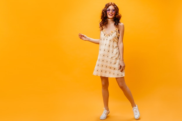 Instantánea en pleno crecimiento de joven delgada en vestido amarillo corto. Mujer pelirroja en gafas de sol posando sobre fondo naranja.