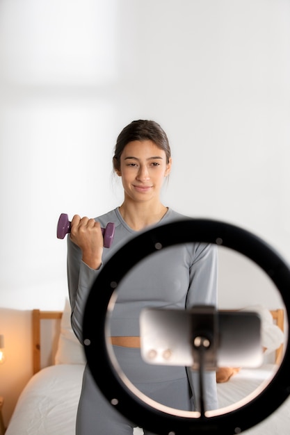 Instructora de fitness femenina grabándose a sí misma levantando pesas en casa con un teléfono inteligente y una luz de anillo