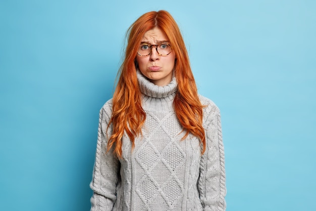 Insultada mujer europea jengibre disgustada tiene expresión ofendida frunce los labios y quiere llorar vestida con un suéter gris lleva gafas.