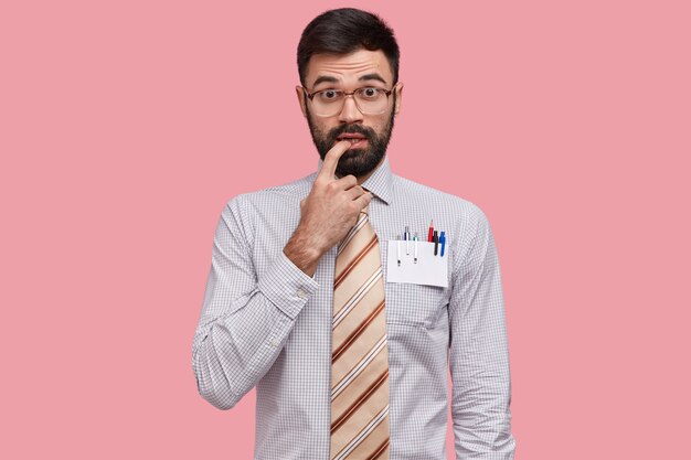 Intelectual indeciso mantiene el dedo cerca de la boca, tiene barba espesa, viste camisa formal y corbata, usa anteojos grandes