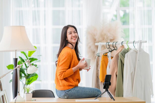 Inteligente, atractiva, sonriente, propietaria de una empresa asiática, empresaria, sostenga la mano, una taza de café, retrato, mirando a la cámara con una mujer confiada sentada en la mesa en el estudio de su casa