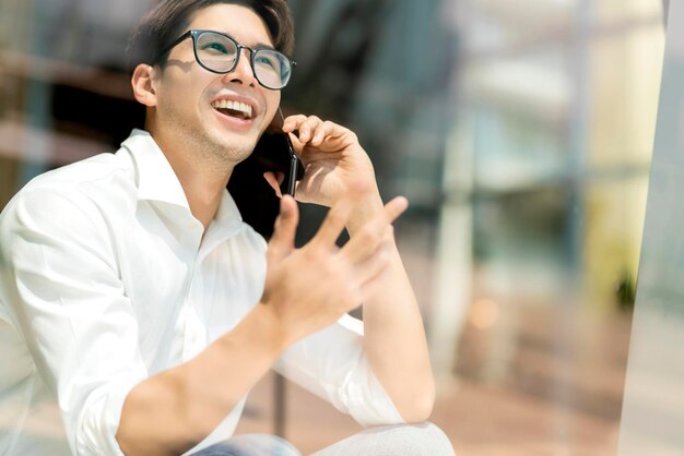 Inteligente, atractivo, positivo, hombre de negocios asiático con anteojos, trabajo remoto, nuevo estilo de vida normal, uso manual, conferencia de teléfonos inteligentes, videollamada en vivo, reunión, ideas de negocios remotas, tomadas a través de la ventana