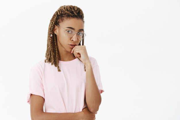 Inteligente, creativa y de aspecto serio joven jefa con rastas en camiseta rosa y gafas con la cabeza apoyada en el puño mirando