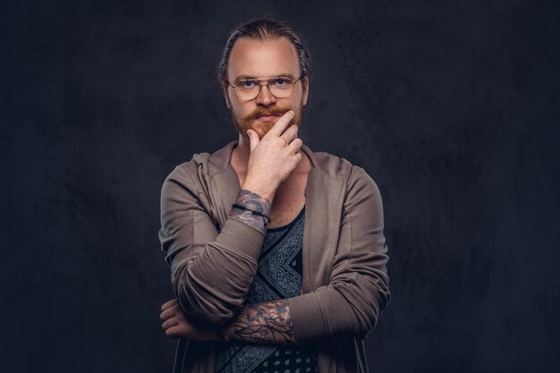 Inteligente hipster pelirrojo pensativo con barba completa y anteojos vestidos con ropa informal, posa con la mano en la barbilla en un estudio. Aislado en el fondo oscuro.