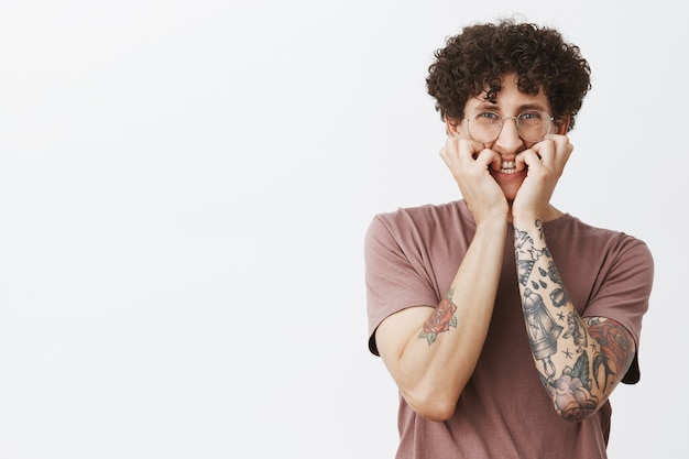 Foto gratuita intenso, asustado y tímido, guapo chico europeo divertido con bigote, cabello rizado y tatuajes, mordiéndose las uñas y frunciendo el ceño, sintiéndose asustado por una película de terror en la televisión.