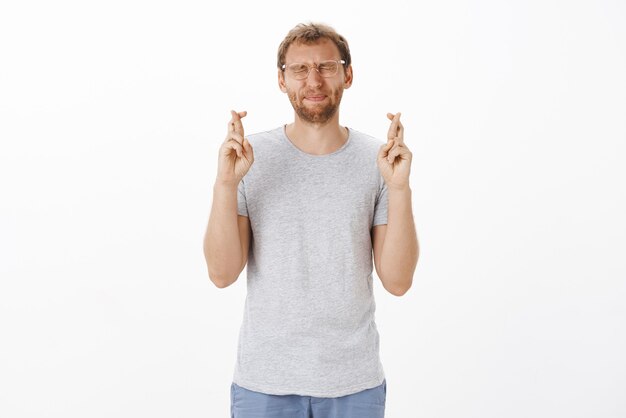 Intenso macho adulto lindo preocupado con cerdas en vasos cerrando los ojos frunciendo los labios y cruzando los dedos para tener buena suerte mientras espera que el plan funcione