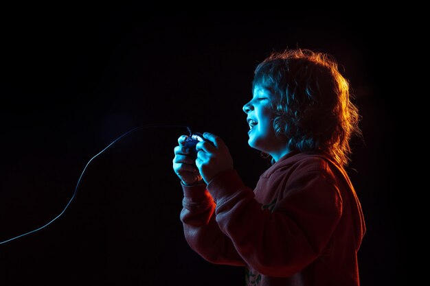 Intenté jugar videojuegos. Retrato de niño caucásico en pared oscura en luz de neón. Preciosa modelo rizada. Concepto de emociones humanas, expresión facial, ventas, publicidad, tecnología moderna, gadgets.