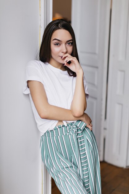 Interesada chica de pelo castaño de buen humor posando en casa. Mujer morena alegre en camiseta escalofriante en su apartamento en fin de semana.