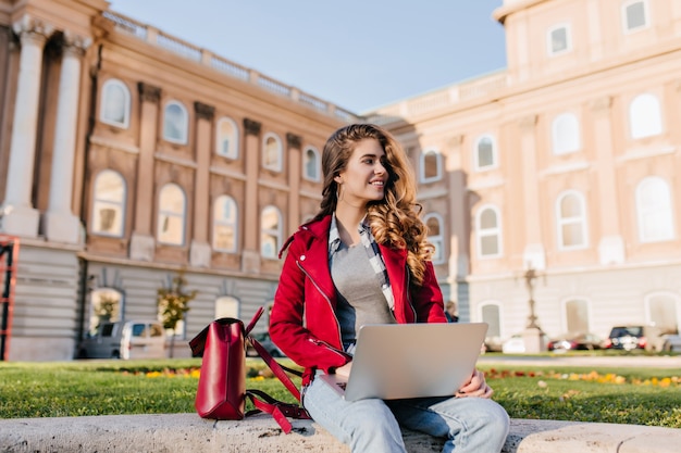 Interesada chica de pelo oscuro viste ropa informal relajándose en el parque cerca de la universidad y usando una computadora portátil