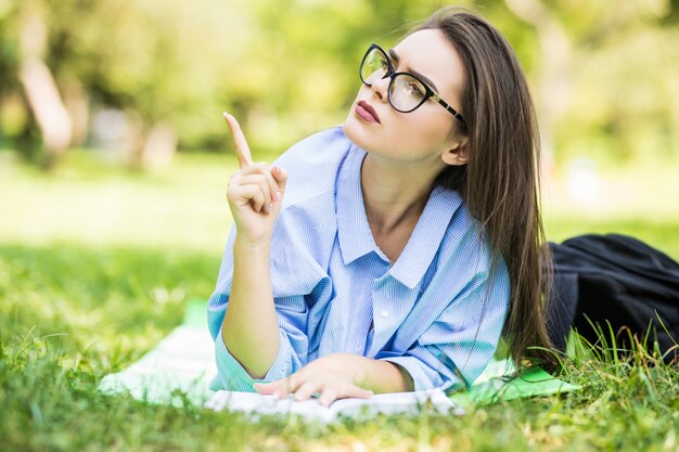 Interesada jovencita tumbado sobre la hierba en el parque con lápiz y cuaderno