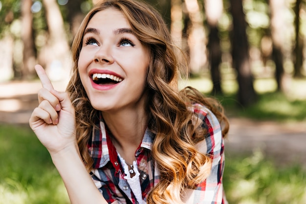 Interesada mujer caucásica de ojos azules mirando hacia arriba con una sonrisa. Chica de moda en camisa a cuadros expresando felicidad en el parque.