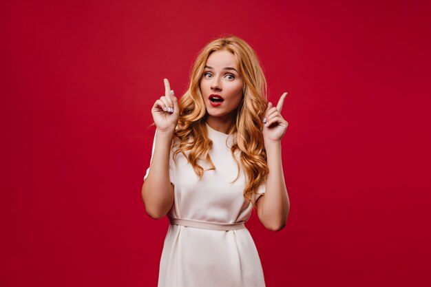 Interesada mujer joven con pelo largo ondulado posando con la boca abierta. Chica elegante elegante en vestido blanco de pie en la pared roja.