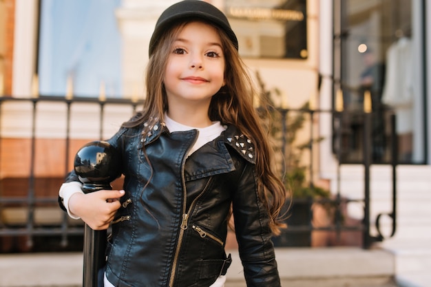 Interesada niña delgada con cabello largo oscuro posando frente al edificio de la escuela junto al pilar de hierro.