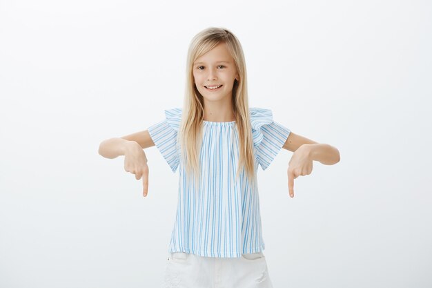 Interesada niña feliz con cabello rubio, apuntando hacia abajo con los dedos índices y sonriendo ampliamente, segura de sí misma y relajada sobre la pared gris