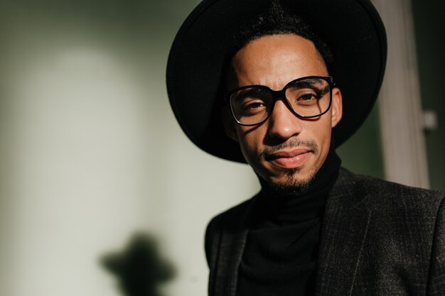 Interesado hombre hermoso con ojos negros mirando. Foto interior de chico africano bien vestido de moda con gafas.