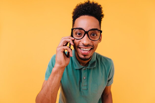 Interesado hombre de ojos oscuros con gafas hablando por teléfono. Chico africano feliz viste ropa verde posando con smartphone.