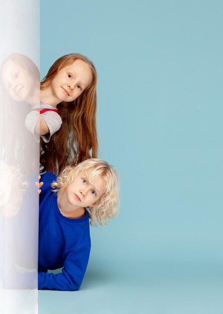 Interesar. Niños felices aislados sobre fondo azul de estudio. Mira feliz, alegre. Copyspace para el anuncio. Infancia, educación, emociones, concepto de expresión facial. Asomándose desde detrás de la pared.