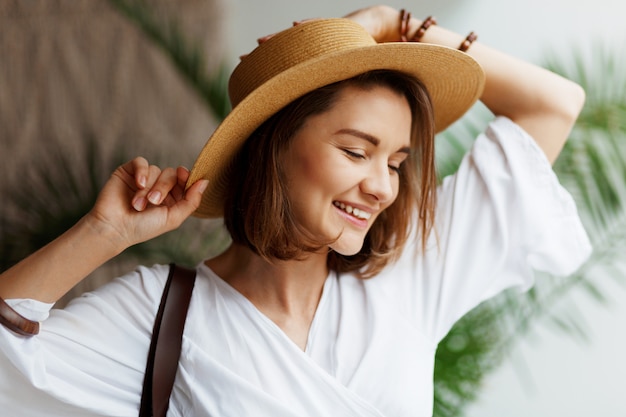 Interior cerca retrato de mujer bonita elegante con sombrero de paja y vestido blanco posando en casa