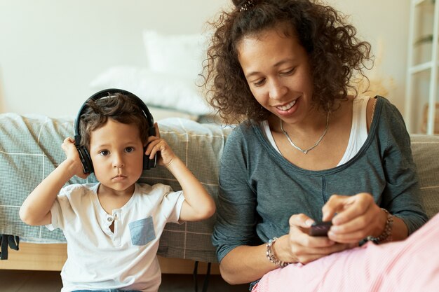 Interior retrato de alegre joven hispana sosteniendo teléfono móvil tocando pistas de música para su adorable hijo que escucha canciones a través de auriculares inalámbricos