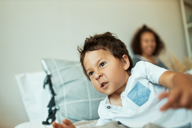 Interior retrato de obstinado niño de 3 años con piel oscura acostado en la cama con la madre, actuando travieso, no quiere dormir