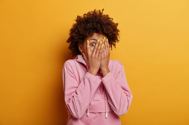 Intrigada curiosa mujer de piel oscura se asoma a través de los dedos, se cubre la cara con las palmas, se esconde, teme algo, tiene los ojos saltones, se viste con una sudadera informal, posa en el interior sobre una pared amarilla