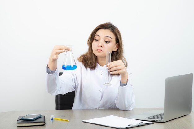 Investigador de laboratorio femenino sentado en el escritorio y sosteniendo un frasco de vidrio médico con líquido azul.