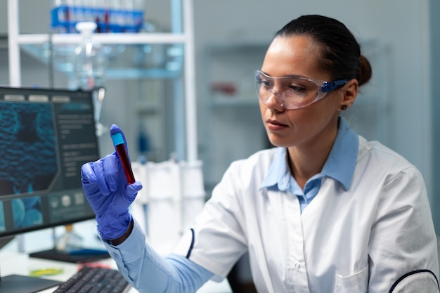 Investigador médico bioquímico analizando vacutainer transparente con sangre trabajando en el experimento de microbiología en el laboratorio del hospital. Mujer bióloga desarrollando vacunas médicas utilizando biotecnología