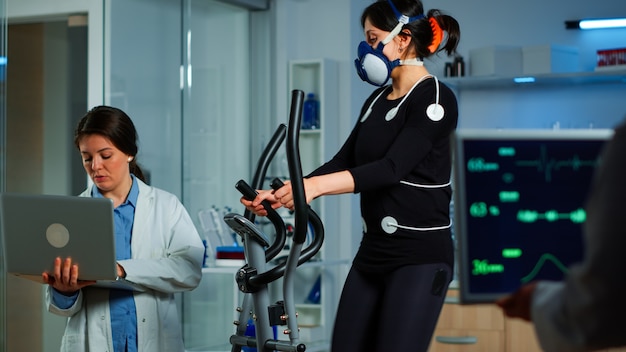 Investigador médico usando una computadora portátil mientras mide la resistencia del deportista usando sensores corporales, electrodos y una máscara que mide el ritmo cardíaco
