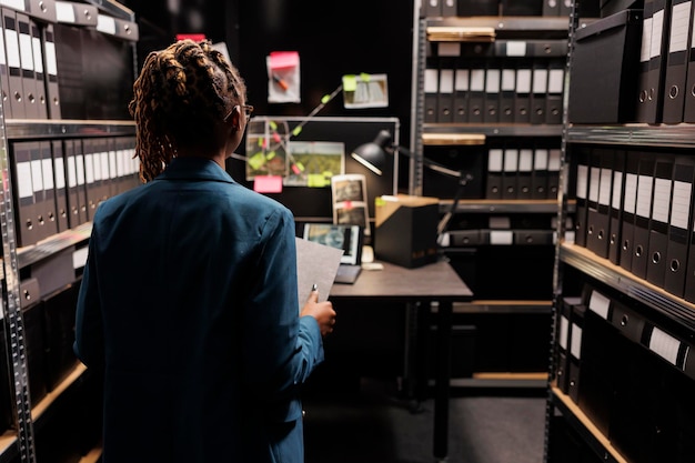 Foto gratuita investigador de la policía que tiene un archivo de asuntos criminales y analiza la junta de evidencia en el cargo tarde. detective afroamericana de las fuerzas del orden estudiando pistas, trabajando horas extras por la noche