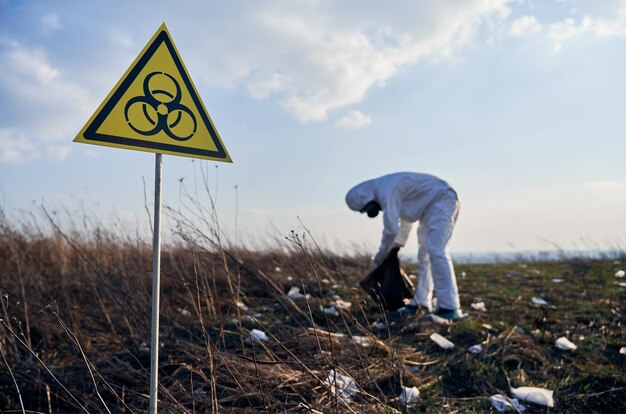 Investigador en traje protector recogiendo basura plástica en una bolsa de basura negra al aire libre en un día soleado