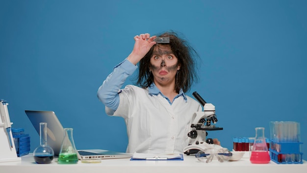 Foto gratuita una investigadora loca y tonta usando un microscopio en el escritorio, actuando loca y loca con el pelo loco. tonto científico tonto trabajando en química, siendo gracioso con la cara sucia después de la explosión de humo.