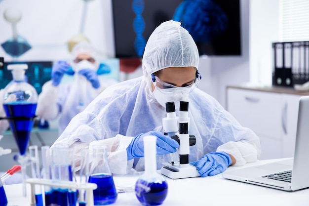 Investigadora mirando a través de un microscopio en el laboratorio de investigación. Fumar líquido azul en tubos de ensayo.