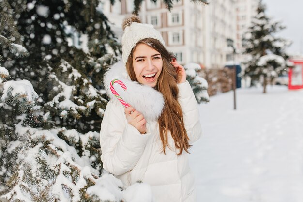 Invierno congelado de divertida mujer asombrosa que se divierte con lollypop rosa en la calle. Joven mujer alegre disfrutando de nevar en chaqueta, gorro de punto, expresando positividad. Delicioso y dulce invierno.